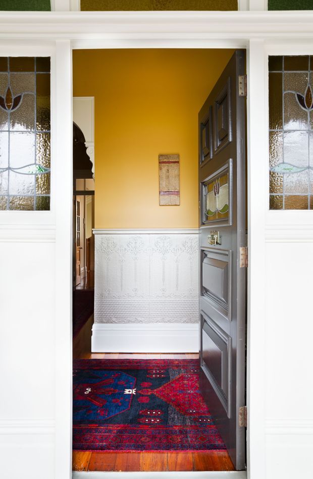 A richly detailed traditional entryway viewed from outside an open front door. The entrance features white decorative wall panelling and trim, ornate stained-glass sidelights, a dark grey raised-panel timber door, a mustard-gold interior wall, polished timber flooring, and a deep red and navy patterned runner rug. A glimpse of an adjoining hall is visible beyond.