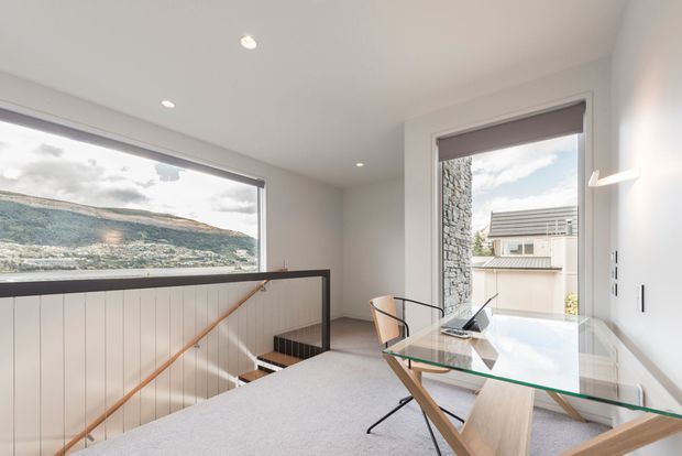 Bright contemporary home office or study positioned beside a stair landing, with a glass-top timber desk, laptop, pale upholstered chair, broad windows, and a partially visible stairwell with white vertical panelling and timber handrail. The room has white walls and ceiling, recessed downlights, carpeted flooring, and roller blinds. Views through the windows show a hilly landscape and neighbouring building, while the interior remains clean, light, and minimalist.