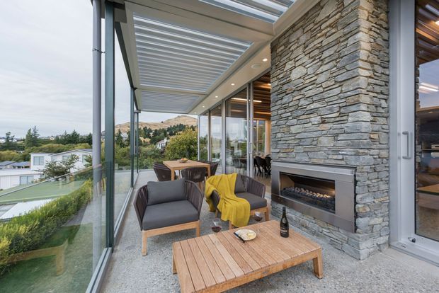 A contemporary covered outdoor lounge adjoining a glazed home, with two upholstered armchairs, a yellow throw, and a low timber coffee table arranged around a linear built-in fireplace. A substantial stacked-stone feature wall, exposed timber ceiling slats, full-height glazing, and a louvred pergola define the space, with landscaped residential surroundings visible beyond.
