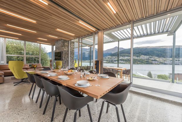 A refined open-plan dining and living area with a long timber dining table set for a meal, sculptural dark dining chairs, and a yellow lounge chair. The room features a timber slatted ceiling with integrated linear lighting, polished concrete or terrazzo-style flooring, a stone-clad fireplace or chimney feature, and extensive floor-to-ceiling glazing. A covered outdoor terrace and a lake with mountainous surroundings are visible beyond the glass.