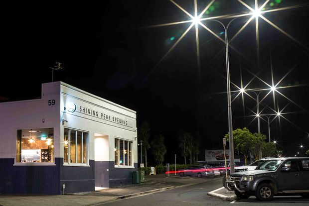 Night-time exterior photograph of a small corner brewery or hospitality venue marked with the address number 59. The white and dark navy facade has tall windows, a glazed entrance, and signage reading “Shining Peak Brewing.” Bright contemporary streetlights illuminate the forecourt and adjacent parking area, with a parked utility vehicle in the foreground.