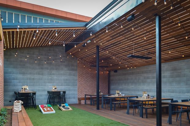 A covered outdoor courtyard dining and recreation area with dark timber slatted pergola ceilings, string lights, steel posts, concrete block and brick walls, picnic-style tables, high bar tables, and artificial grass with cornhole boards. The space has a contemporary industrial hospitality feel and appears connected to a larger building.