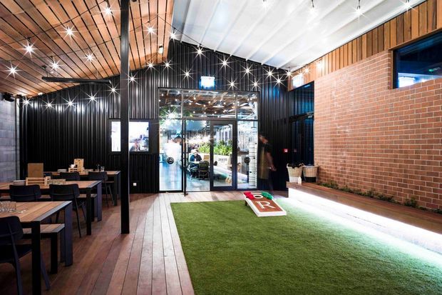 A contemporary commercial interior, likely a hospitality or recreational venue, with a long artificial-grass activity area and cornhole boards in the foreground. The space combines timber plank flooring and a timber-lined ceiling with black corrugated metal walls, exposed dark steel posts, a red brick feature wall, extensive glazed doors, and café-style dining tables and chairs. Bright linear and point lighting creates a lively, high-contrast atmosphere.