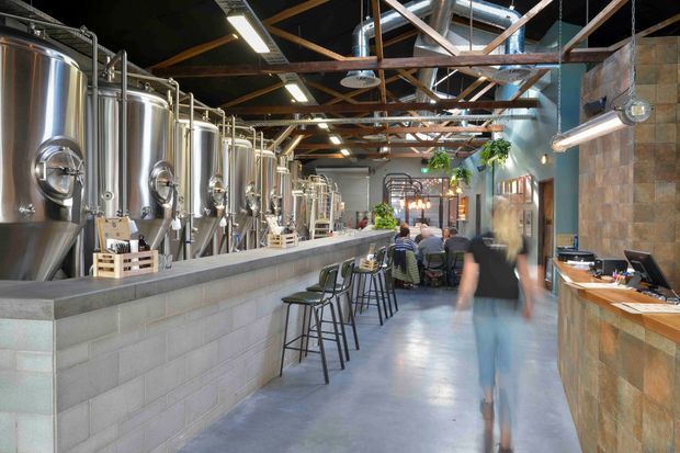 A spacious industrial-style brewery taproom or hospitality venue, with large stainless-steel brewing tanks lining the left side behind a concrete-block service counter. The space has a polished concrete floor, exposed timber roof structure, visible metal ductwork, linear industrial lighting, and a textured masonry bar or reception counter on the right. Bar seating and tables occupy the central aisle, with patrons and greenery near the rear.