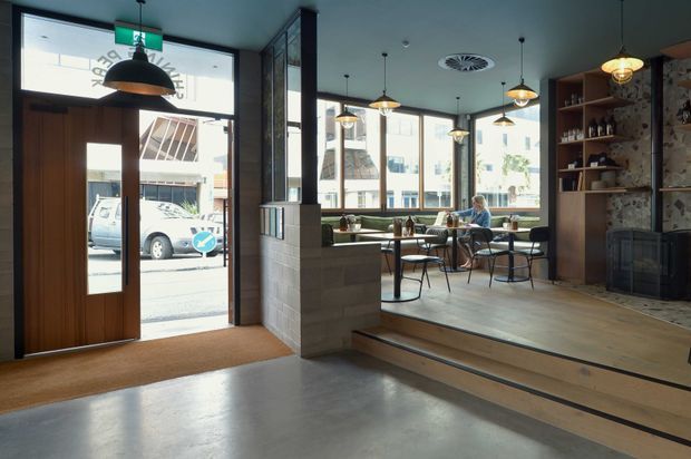 Interior view of a contemporary café or hospitality venue, photographed from the entrance. The space features a polished concrete floor, timber-framed glazed entry, raised timber dining platform, exposed masonry partition, dark timber shelving with bottles and decor, pendant lighting, and large street-facing windows. A fireplace-like dark metal feature is set into a textured feature wall, with café tables and seating arranged near the glazing.