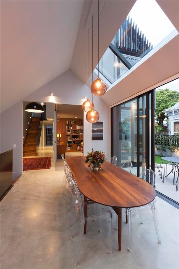 A contemporary dining area with a long rounded timber dining table and transparent acrylic chairs, positioned beside full-height black-framed glazing opening to an outdoor courtyard. Three amber pendant lights hang beneath a high sloping ceiling, while a skylight brings in natural light. A stairway, built-in shelving, and warm timber cabinetry are visible in the adjoining interior.