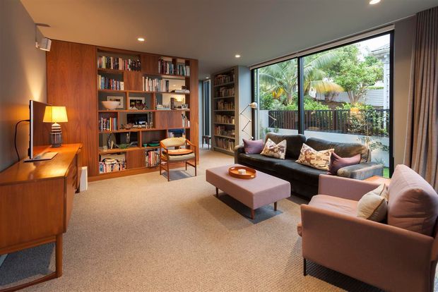 A warm, well-appointed living room with a mid-century-inspired timber entertainment console and extensive built-in bookshelf wall. The room features a dark sofa, two upholstered armchairs, a rectangular ottoman coffee table, carpeted flooring, recessed ceiling lights, floor-to-ceiling curtains, and large glazed doors or windows opening to a leafy outdoor area.