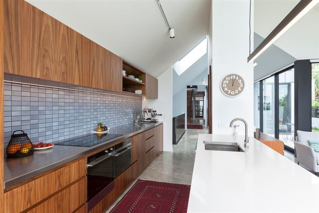 A bright contemporary open-plan kitchen with extensive dark walnut-toned cabinetry, a long grey tiled splashback, dark benchtops, integrated oven and cooktop, and a large white island with a stainless-steel sink and curved tap. Skylights, clerestory glazing and large black-framed windows bring in abundant daylight, with a living area visible beyond the island.