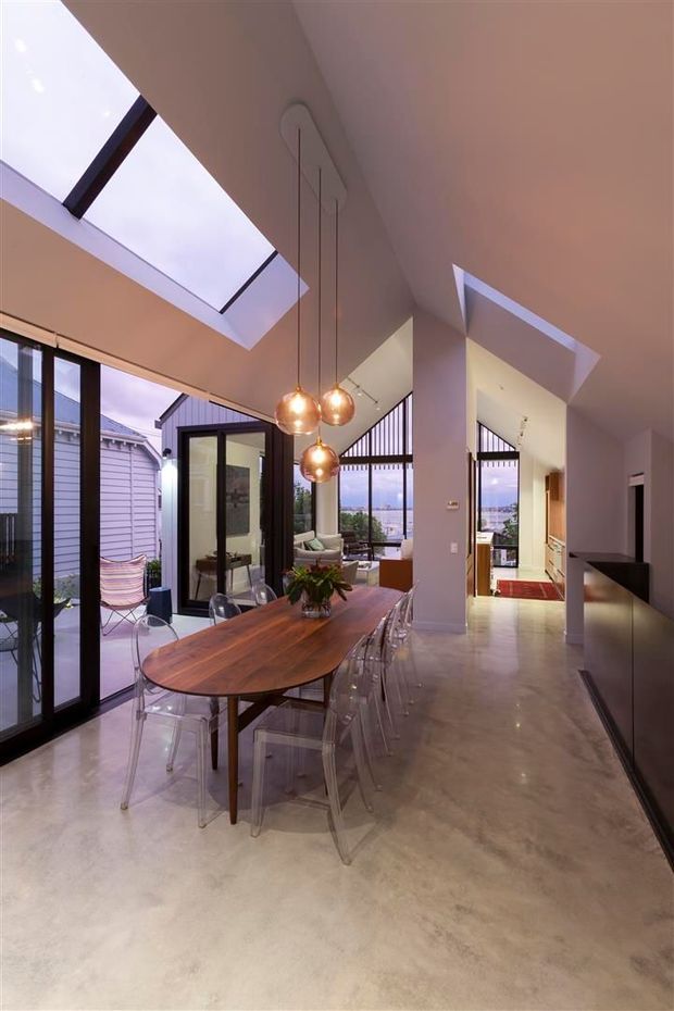Open-plan contemporary dining and living area with a long timber dining table, transparent acrylic chairs, polished concrete flooring, and three suspended amber globe pendant lights. Large black-framed glazed doors, extensive windows, and multiple skylights bring in natural light. A lounge area and kitchen circulation are visible beyond the dining zone, with an outdoor patio immediately alongside.
