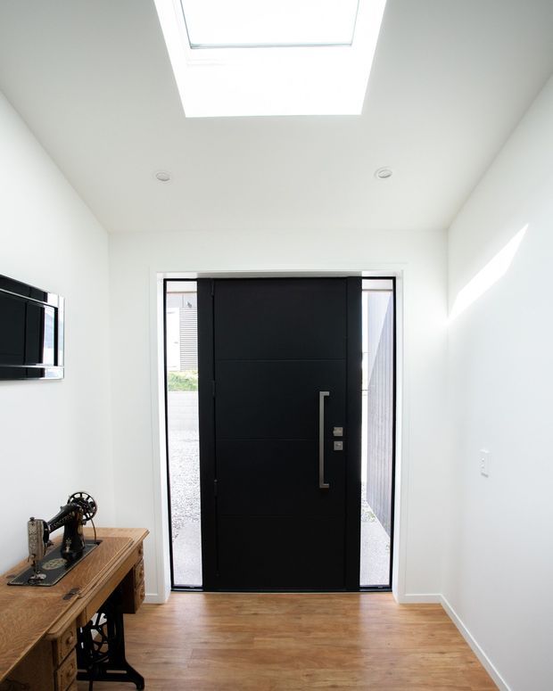 Bright, narrow residential entryway with white walls and ceiling, warm timber flooring, a large matte-black pivot-style front door with slim glazed sidelights, and a roof skylight. A vintage sewing machine on a timber cabinet sits to the side, while sunlight creates a strong diagonal highlight on the wall.