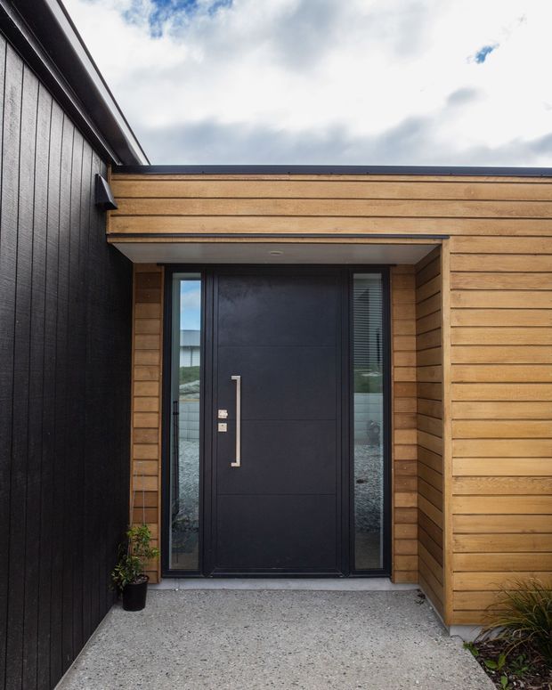 Exterior close-up of a contemporary residential entrance. A matte black front door with a long vertical metal handle is framed by narrow sidelights and recessed beneath a small timber-clad canopy. The surrounding facade combines warm horizontal timber weatherboards with dark vertical cladding, opening onto a textured concrete entry path.