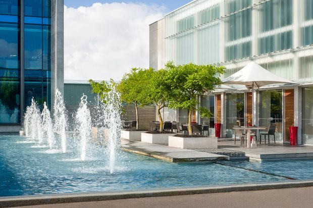 Contemporary commercial building exterior with a large fountain pool in the foreground and a landscaped terrace seating area beneath a white canopy. The facade combines blue-tinted glazing, translucent vertical panels, and metal framing, with outdoor tables and chairs along the water feature.