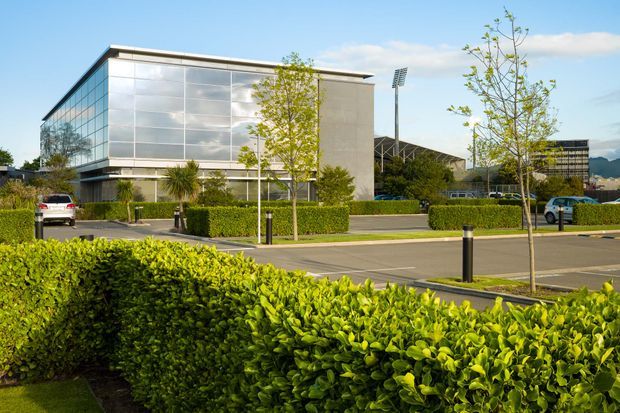 Exterior view of a modern commercial building with a large reflective glass curtain wall, pale concrete or stone side wall, and low flat roof. The building sits within a landscaped commercial or institutional campus with clipped hedges, young trees, paved parking, bollards, and sports floodlights visible behind it.