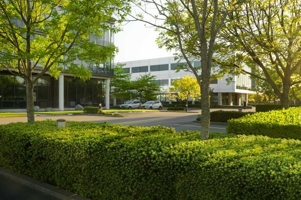 Exterior view of a modern multi-storey commercial building set within landscaped grounds. The white and grey facade features extensive horizontal glazing, projecting volumes, and a recessed glazed entrance, framed by mature trees and neatly clipped hedges.