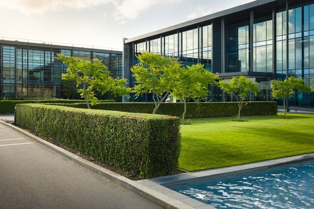 Contemporary commercial building with expansive dark-framed glass façades, landscaped lawns, manicured hedges, small ornamental trees, and a reflective pool along the foreground. The scene has a clean, formal corporate campus character.