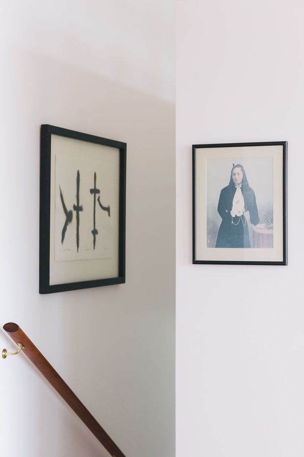 A softly lit interior stairway or landing corner with white walls, two black-framed artworks, and a dark timber handrail with a small brass wall bracket. The composition is minimal and refined, focusing on the artwork and stair detail.