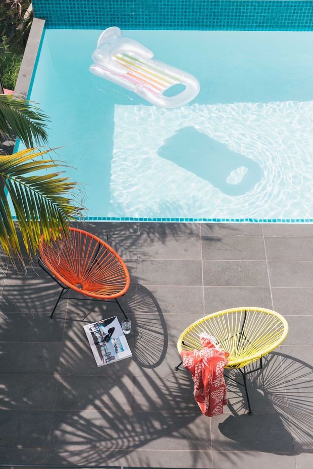 A bright overhead view of a turquoise swimming pool bordered by small blue mosaic tiles and dark grey square paving. Two colourful woven lounge chairs, tropical palm foliage, and an inflatable pool lounger create a relaxed resort-like outdoor setting.