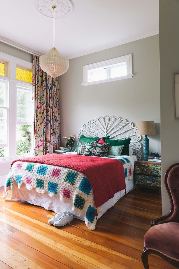 Bright, characterful bedroom with a bed positioned beside tall bay-style windows. The room features polished warm timber floorboards, muted sage-grey walls, white decorative ceiling moulding and window trims, a floral patterned curtain, a white woven fan-shaped bedhead, colourful crocheted and patterned bedding, and a vintage-style painted bedside table. A suspended faceted pendant light and upholstered armchair add decorative detail.