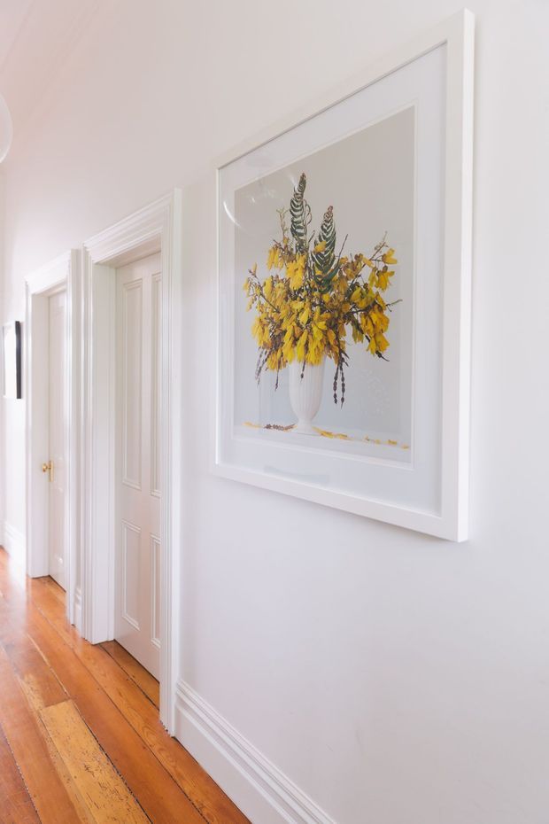 A bright, narrow residential hallway with white painted walls, ornate white panelled door and architrave detailing, decorative wall mouldings, and polished warm timber floorboards. A large white-framed botanical artwork with yellow flowers is the main wall feature. The corridor has a refined traditional character and opens to several rooms along the left side.