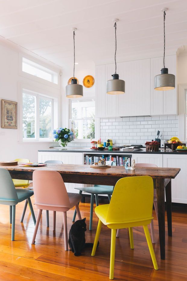 Bright, colourful kitchen-dining space with white vertical wall panelling, white subway-tile splashback, timber flooring, a substantial rustic timber dining table, and three grey pendant lights. A compact kitchen with white cabinetry and dark benchtops sits behind the dining area, with colourful dining chairs, fresh produce, flowers, and open shelving adding character.