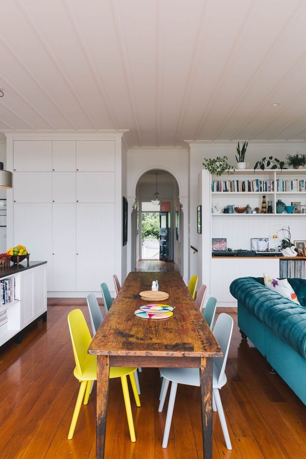 A bright, eclectic open-plan dining and living space. A large rustic timber dining table with colourful mismatched chairs dominates the foreground, while a deep teal sofa sits to the right. White built-in cabinetry and open bookshelves line the walls, with decorative objects and plants on display. The room features warm timber flooring, an arched hallway opening, and a white beadboard-style ceiling with panelled detailing.
