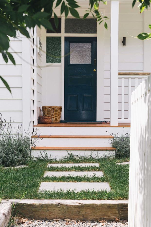 A residential front entry viewed from the garden path, with a dark navy-blue paneled door featuring textured glass sidelights and transom, set within white horizontal weatherboard cladding. The raised porch has white steps with timber nosing, a white picket fence borders the path, and stepping stones cross a lawn with low silvery planting. A woven basket sits beside the doorway. The scene has a clean, bright, welcoming character with traditional cottage styling.