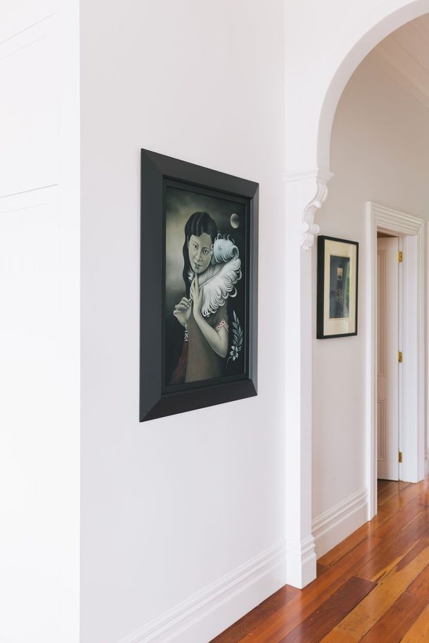 A bright, refined residential hallway with white walls, decorative architraves, ornate moulding, and polished timber floorboards. Framed artwork hangs along the corridor, with a curved arched opening leading to adjoining rooms.