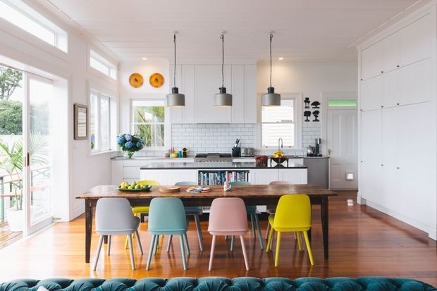 Bright, open-plan kitchen and dining area with white built-in cabinetry, a pale grey subway-tile splashback, timber flooring, and a large rustic timber dining table. Three industrial-style pendant lights hang above the island, while colourful dining chairs and small decorative accents add a playful touch. Tall glazing and a glazed exterior door bring in abundant natural light; a dark teal tufted sofa is partially visible in the foreground.