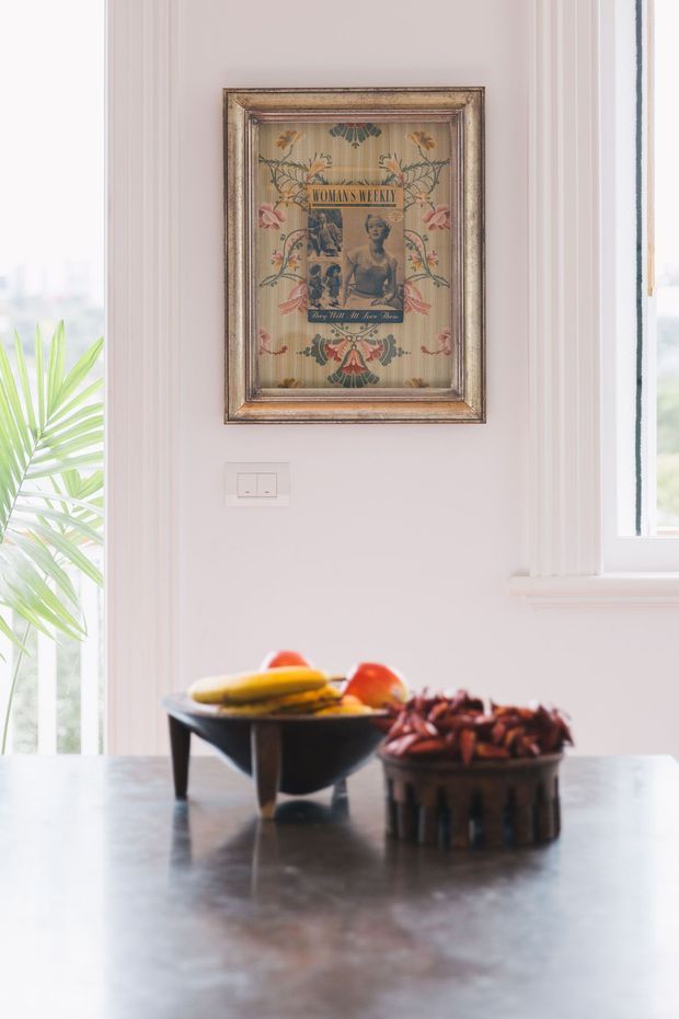 A softly lit interior vignette featuring a dark stone or polished benchtop/table in the foreground, two decorative fruit bowls, pale painted walls with vertical moulded trim, tall white-framed windows, and a vintage framed Woman's Weekly artwork. The scene has a bright, refined, heritage-inspired character with botanical greenery visible near the window.