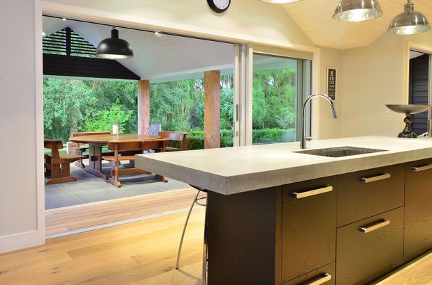 A contemporary kitchen with a large dark-wood island and pale stone benchtop, integrated sink and curved chrome tap. Pendant lights hang above the island, while wide sliding glass doors open to a covered outdoor dining area surrounded by greenery. Timber flooring continues toward the outdoor deck, and the kitchen has a warm, open-plan connection to the exterior.