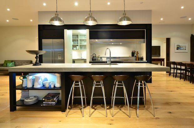 Open-plan kitchen dominated by a large island with a pale stone benchtop, dark cabinetry, integrated shelving, stainless-steel appliances, and four pendant lights. Timber flooring continues into the adjacent dining and living areas, creating a warm, contemporary entertaining space.