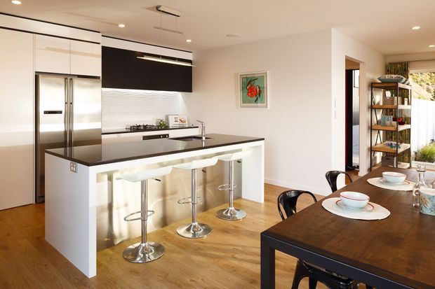 Bright open-plan kitchen and dining area with white full-height cabinetry, stainless-steel refrigerator, dark overhead cabinetry, a white island with dark benchtop and integrated breakfast bar, and three chrome stools. A dark timber dining table occupies the foreground beside open shelving and a doorway. The space has warm timber flooring, recessed lighting, and a white textured splashback.