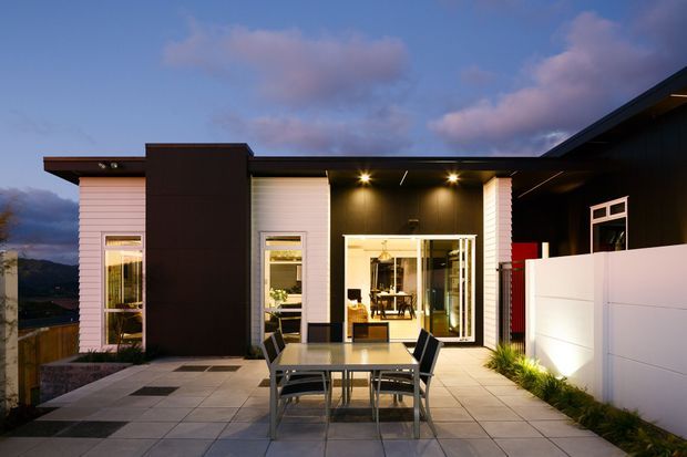 Contemporary single-storey home exterior viewed from a paved outdoor entertaining terrace at dusk. The facade combines white horizontal cladding with dark charcoal feature walls and roofing, large glazed sliding doors, recessed downlights, and a white privacy boundary wall. A dining table and chairs occupy the terrace, with distant hills visible beyond the property.