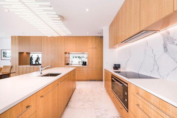 Bright contemporary kitchen with extensive warm timber cabinetry, a large white island, integrated appliances, and a dramatic white marble-look splashback. Linear sculptural ceiling lighting and a polished light floor create a refined, spacious feel.