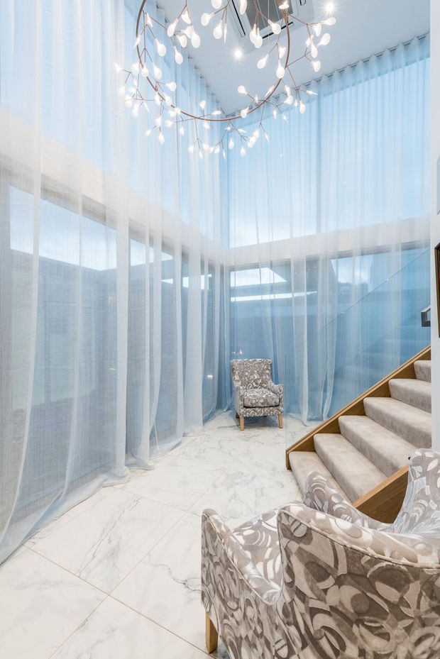 Bright double-height interior with sheer pale blue curtains draped across extensive glazing and walls, a sculptural branching pendant light, white marble-look tiled flooring, patterned upholstered armchairs, and a carpeted staircase with timber edging and glass balustrade. The space feels airy, elegant, and refined.