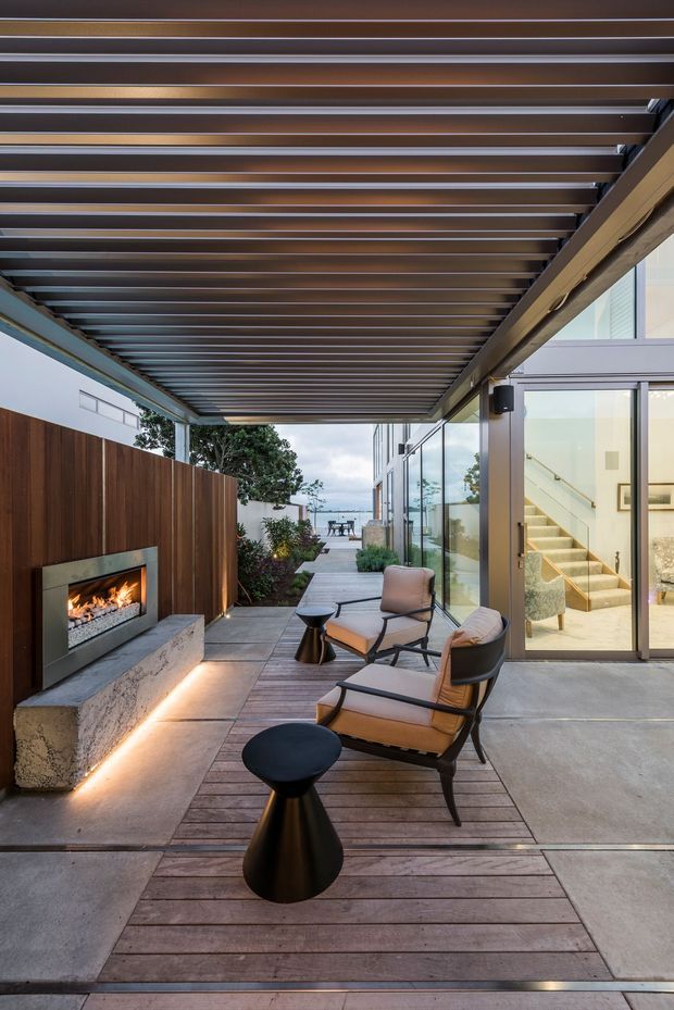 A contemporary covered outdoor terrace beside a glass-walled residence. The space features a slatted pergola roof, timber decking, concrete paving, a built-in linear fireplace set into a dark timber-clad wall, and two lounge chairs with small black side tables. Glass doors reveal a bright interior stairway, while a landscaped walkway leads toward a distant coastal-looking view.