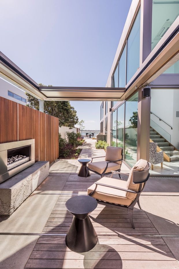 A bright contemporary outdoor courtyard terrace between modern glass-fronted building wings. The space features timber decking and paving, a vertical timber privacy fence, a built-in outdoor fireplace with stone or concrete surround, a slim pergola or retractable canopy, and mid-century-style lounge chairs with small dark side tables. Large glazed doors reveal an interior stairway and connect the terrace to the building.