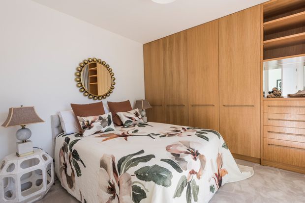 A warm, contemporary bedroom with a floral-patterned bedspread, upholstered bed, bedside tables and lamps, a decorative round mirror, and extensive built-in timber wardrobe cabinetry. An open shelving and drawer unit is visible at the right, with soft neutral finishes and layered natural wood tones.