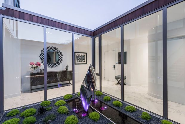 Contemporary courtyard atrium viewed from outside, with floor-to-ceiling glazing framed in dark metal, white marble-look flooring, a reflecting water feature, sculptural dark artwork with purple lighting, and clipped green planting in a gravel garden. A refined interior hall and console are visible through the glass.