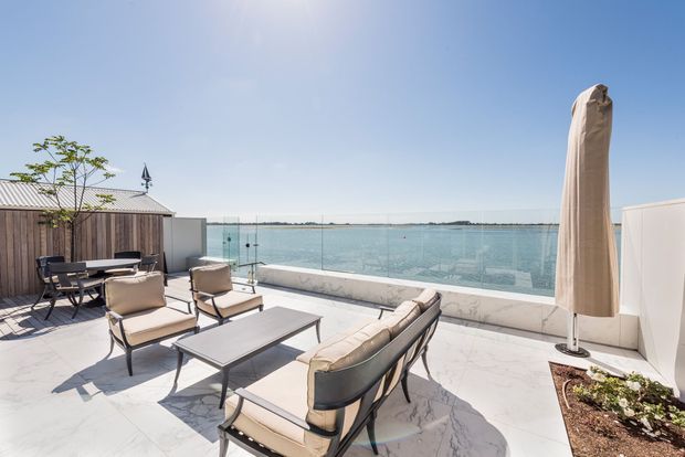 A bright rooftop terrace with a large outdoor seating arrangement overlooking calm water. The terrace features pale marble-look paving, glass balustrades, a timber privacy screen, contemporary lounge furniture, and a closed outdoor umbrella, creating an airy waterfront entertaining space.
