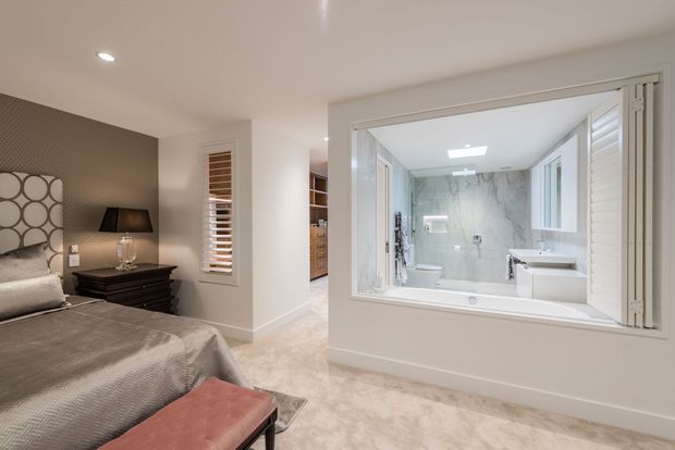 A refined bedroom with a large upholstered bed, mirrored geometric headboard, dark bedside furniture, and a blush upholstered bench. A wide internal window with louvred shutters looks into a bright marble-finished bathroom containing a bathtub, toilet, and double vanity. Neutral tones and soft carpet create a calm, polished atmosphere.