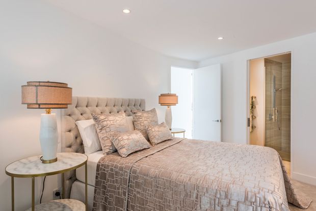 A bright, softly styled bedroom with a tufted upholstered bed, layered patterned bedding and cushions, matching bedside lamps, and a marble-topped round bedside table. A partially open door reveals a modern tiled shower bathroom directly connected to the bedroom.