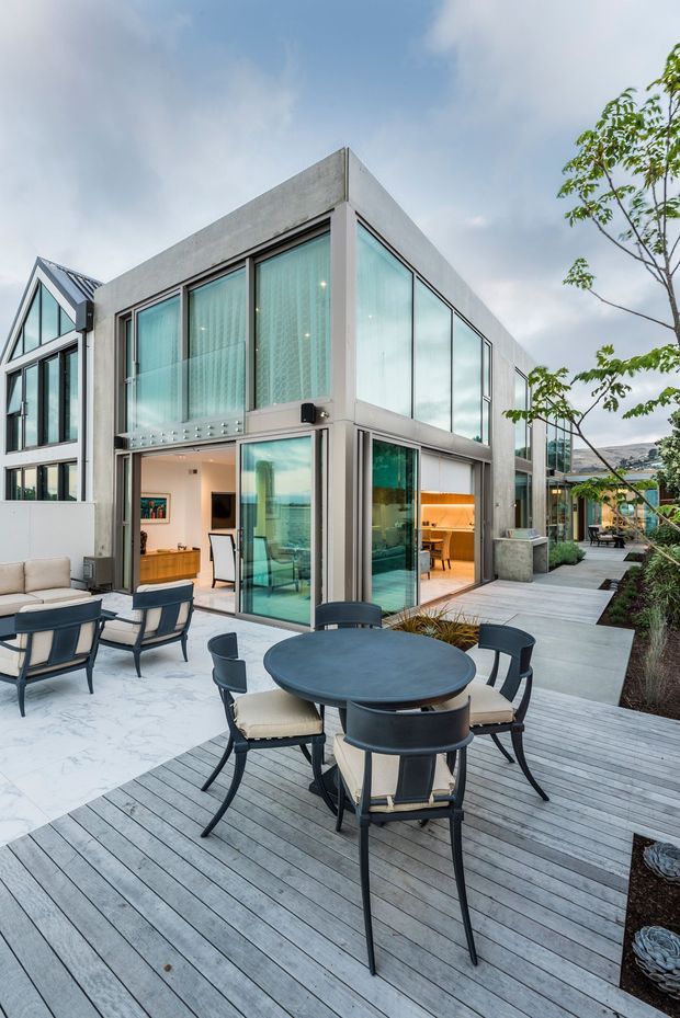 A contemporary two-storey home with a minimalist concrete and extensive glazed facade, opening onto a spacious outdoor entertaining terrace. The terrace combines grey timber decking with a pale paved area, outdoor dining furniture, lounge seating and landscaped garden beds. Large glass doors reveal a bright interior and a warm timber-toned kitchen or dining area. The building has strong rectilinear geometry, floor-to-ceiling glazing and a flat roof, with a neighbouring gabled structure visible at the left.