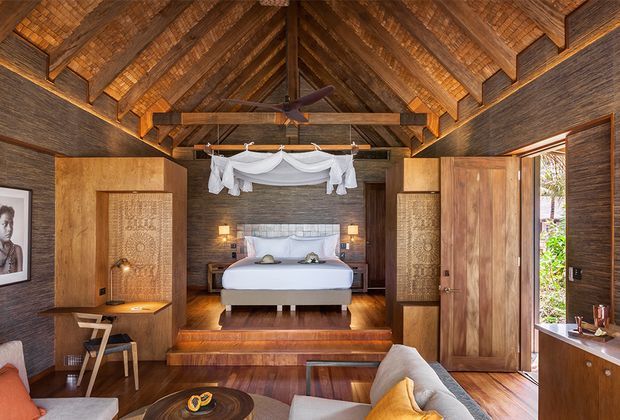 Warm, richly detailed bedroom with a raised sleeping platform, exposed timber truss ceiling, dark textured wall finish, timber flooring, and large timber wardrobe or partition doors. A canopied bed sits centrally with bedside lighting, while a lounge area and low coffee table occupy the foreground. Open doors provide views toward lush greenery outside.