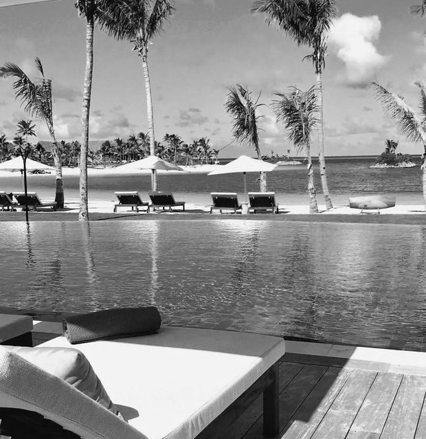 Black-and-white photograph of a resort-style poolside terrace overlooking a tropical beach or lagoon. A large pool occupies the foreground, with a cushioned sun lounger and dark timber decking at the edge. Palm trees, white sun umbrellas, additional loungers, sandy shoreline and calm water create a tropical waterfront setting.