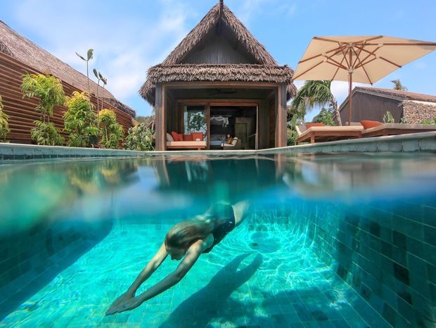 A split-level view across a vivid turquoise swimming pool, with an underwater swimmer in the foreground and a tropical resort-style pavilion beyond. The pavilion has a steep thatched roof, dark timber framing, and an open glazed living area; surrounding structures feature timber cladding and thatched roofing. A poolside lounge with a large beige umbrella and sun loungers sits to the right, with lush tropical planting and palm trees in the background. Bright, sunny, relaxed holiday atmosphere.