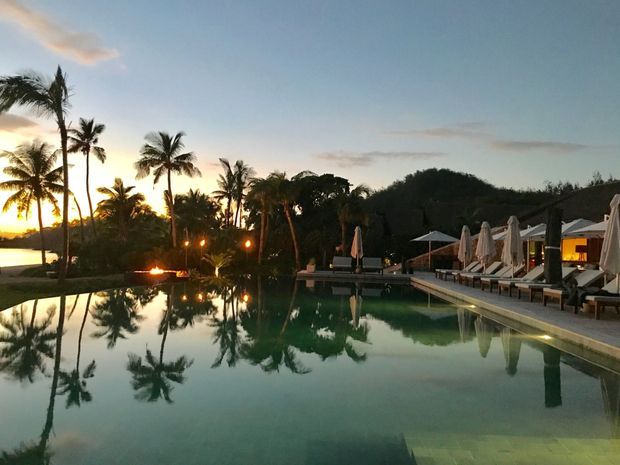 A tranquil resort-style swimming pool at sunset, bordered by palm trees and dense tropical landscaping. The pool reflects the palms and evening sky, while a paved pool deck on the right contains rows of loungers and closed umbrellas beside a low pavilion or poolside building.