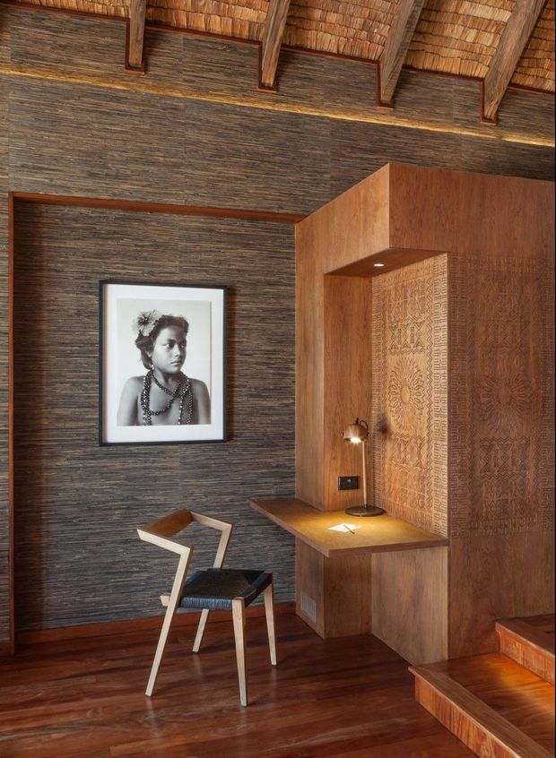 A warm, richly textured study nook or workspace with a compact built-in timber desk, reading lamp, sculptural chair, framed black-and-white portrait, and integrated timber wall panels. The space features dark horizontal textured wall cladding, polished dark timber flooring, carved decorative timber screens, and a dramatic exposed pitched ceiling with natural reed or thatch infill and timber rafters. Steps rise beside the built-in joinery.