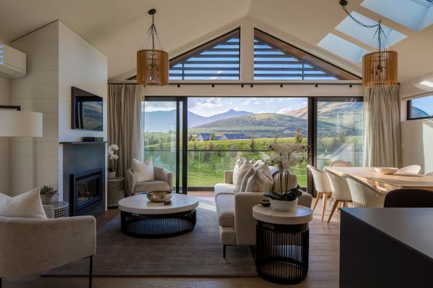 A bright, refined open-plan living and dining area with a dramatic glazed gable end overlooking a green valley and mountain landscape. The room features a fireplace with built-in cabinetry, contemporary upholstered seating, round coffee tables, pendant lights, timber-toned flooring, and a dining area beside expansive glazing. High-level triangular windows and skylights add daylight and emphasize the pitched roof form.