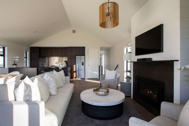 Bright open-plan living room with a large white sectional sofa, round coffee table, armchairs, and a black fireplace beneath a wall-mounted television. The space opens to a modern kitchen with dark timber cabinetry and stainless-steel appliances. White horizontal wall panelling, a vaulted ceiling, timber pendant light, and large windows create a warm contemporary coastal feel.